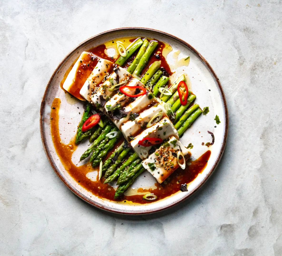 Auf einem Teller zubereiteter Tofu auf grünem Spargel, garniert mit Frühlingszwiebeln, Chilischoten und Sesam,  mit brauner Sauce